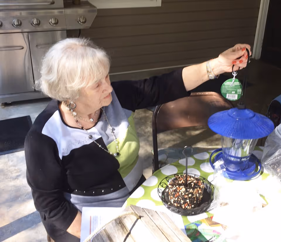 An elderly woman with white hair sitting at a table outdoors, holding a blue bird feeder with a tag attached. The table has a green and white polka dot tablecloth and a round bird seed cake on a black stand. A stainless steel grill is visible in the background.