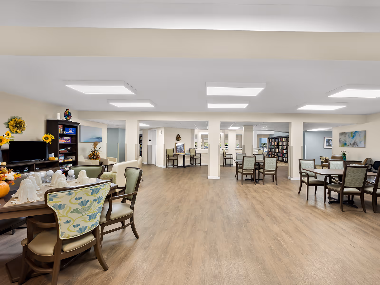 A spacious, well-lit common area in a senior living facility with multiple tables and chairs arranged for socializing or activities. The room features light wood flooring, white walls, and ceiling lights. There is a TV on a stand with shelves holding games and decorations, and some floral decorations are visible on the tables and shelves. In the background, there are bookshelves and additional seating areas.
