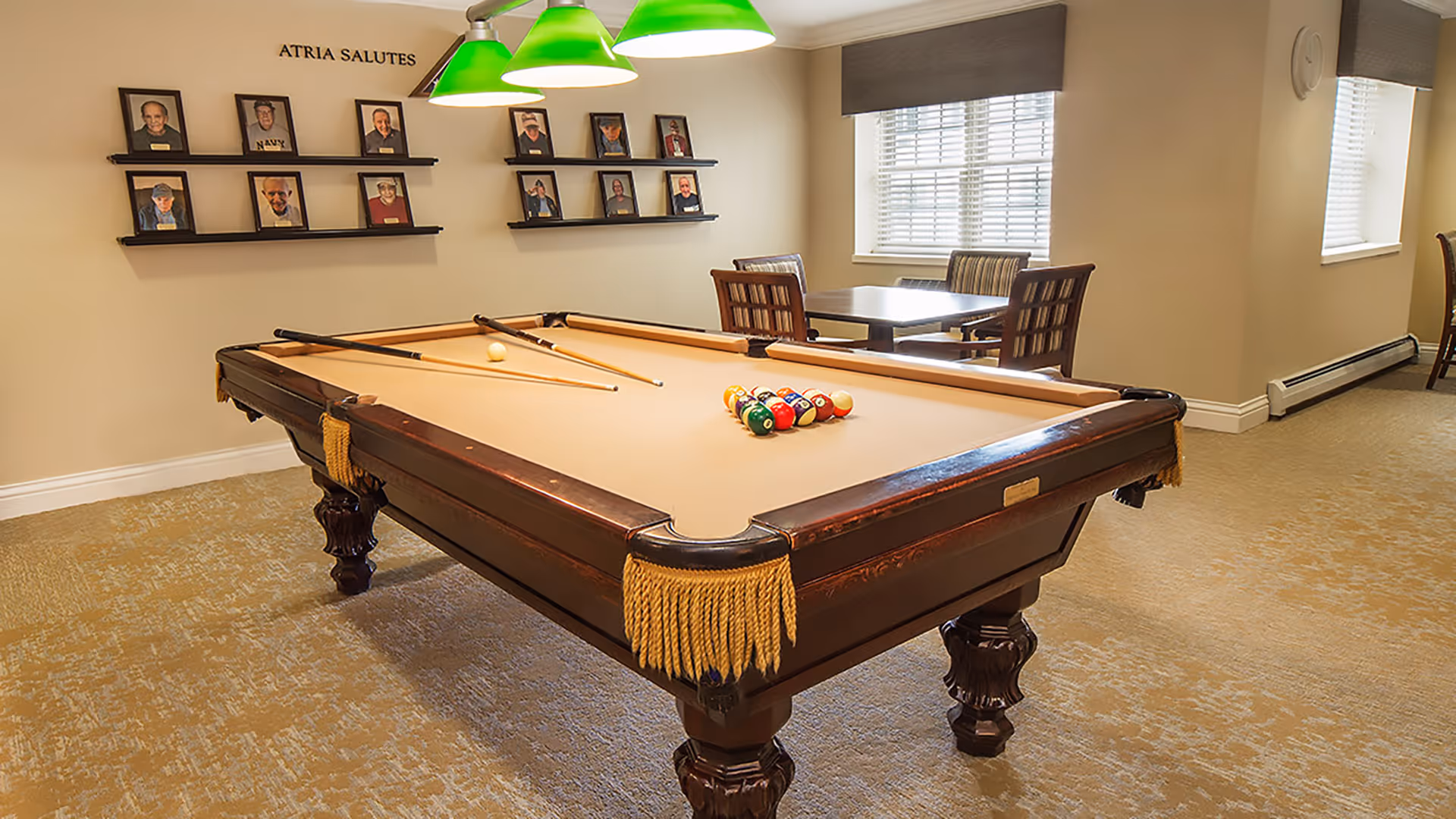 Interior recreation room with a pool table under green hanging lights, framed photos on the wall, and a table and chairs by the window.
