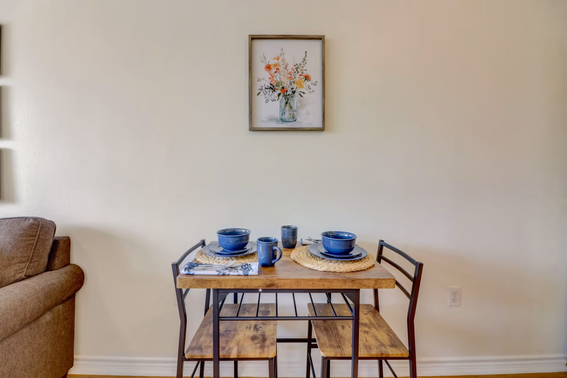 A small wooden dining table set for two with blue dishes, bowls, and mugs on woven placemats. The table has two matching wooden chairs with black metal frames. A framed floral painting hangs on the beige wall above the table. Part of a brown sofa is visible on the left side.