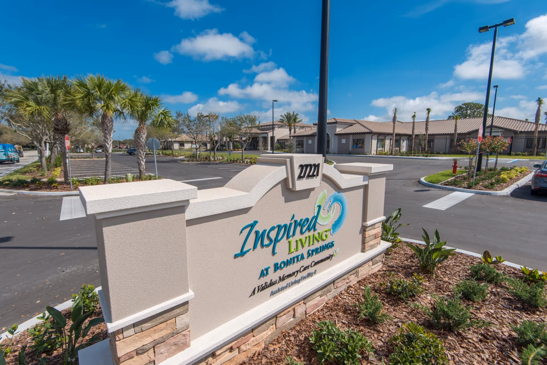 Outdoor view of the entrance sign for Inspired Living Bonita Springs, an assisted living facility. The sign is beige with stone accents and displays the facility name and address 27221. Surrounding the sign are landscaped plants and palm trees, with the facility building and parking lot visible in the background under a blue sky with scattered clouds.