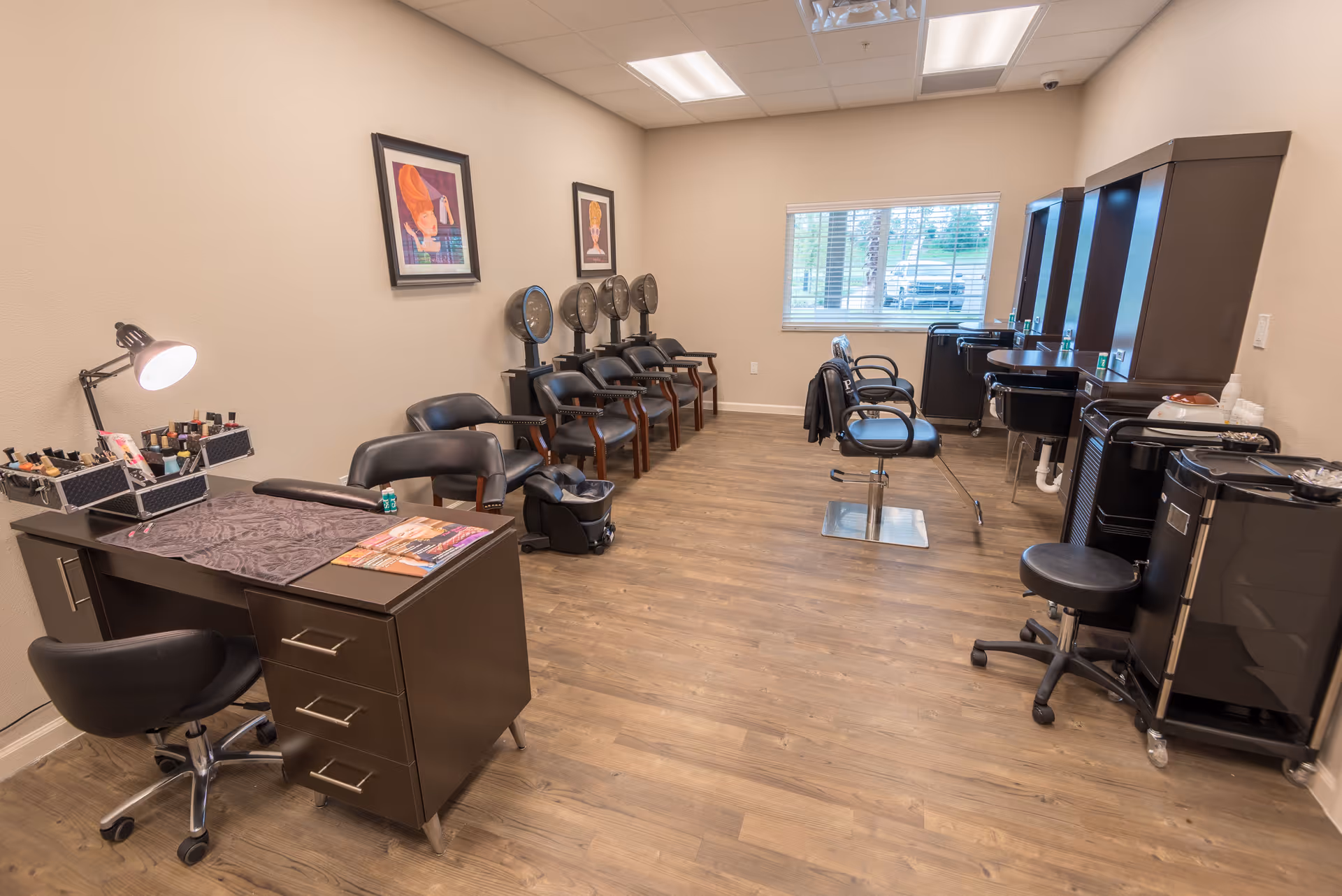 A beauty salon room with wooden flooring and beige walls. The room contains a manicure table with a lamp and nail polish, several black salon chairs, hair drying stations with hooded dryers, and styling stations with mirrors and chairs. Two framed artworks hang on the wall, and a window with blinds lets in natural light.