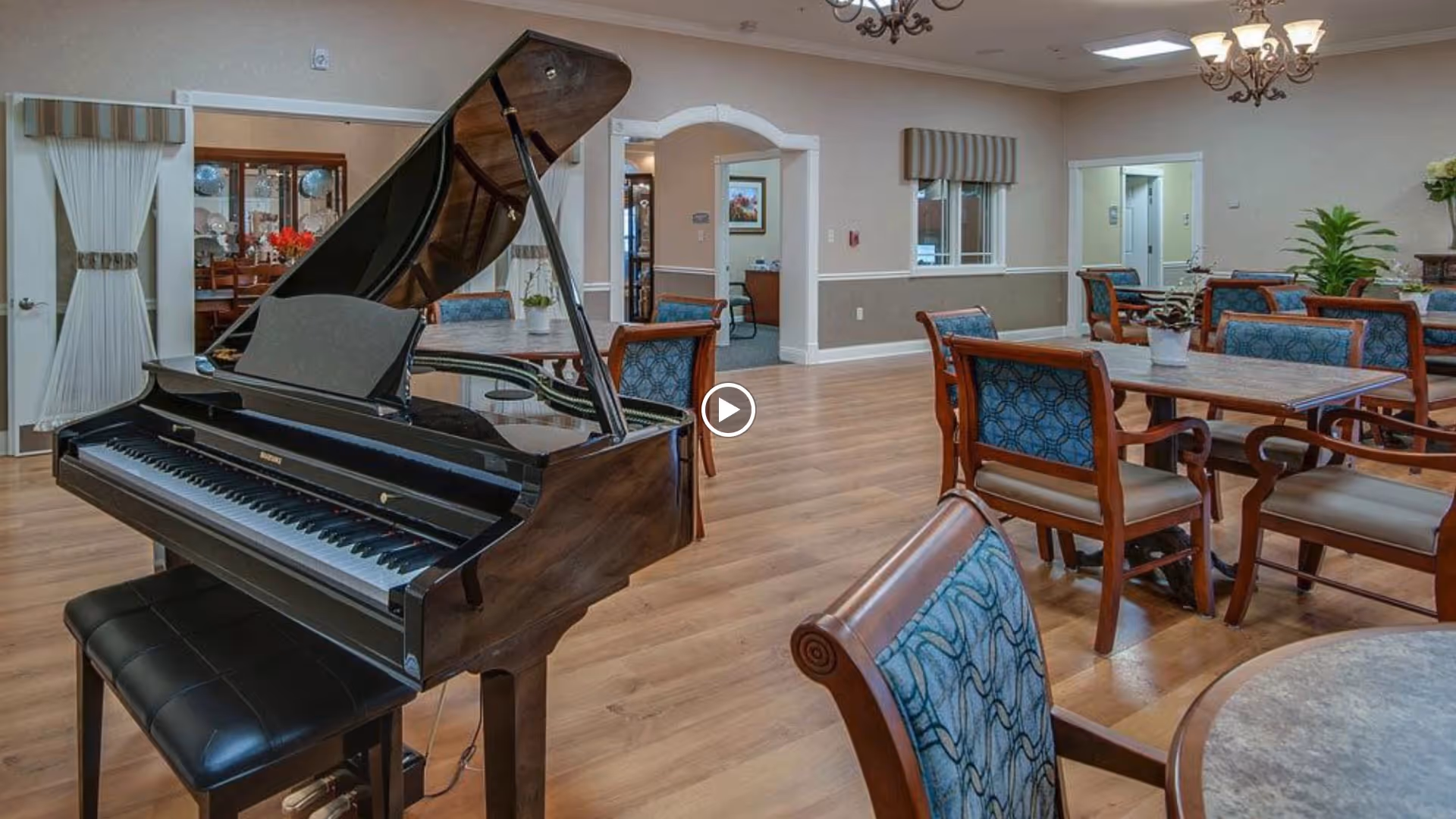 A spacious senior living common area featuring a grand piano with an open lid and a black cushioned bench. The room has wooden flooring and several tables with blue patterned cushioned chairs arranged around them. The walls are light-colored with white trim, and there are chandeliers hanging from the ceiling. In the background, there are doorways leading to other rooms and a window with a striped valance.
