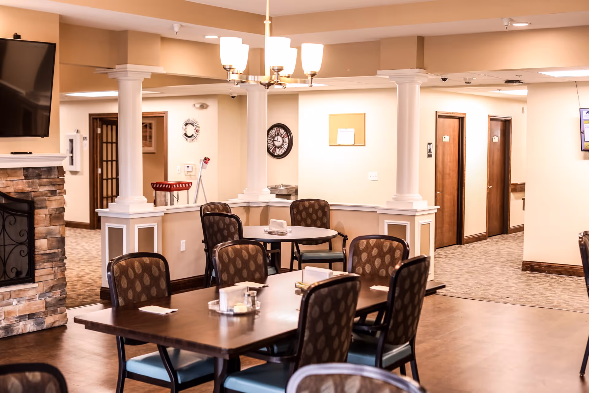 Dining area with tables and chairs, decorative columns, and a fireplace in a senior living facility.