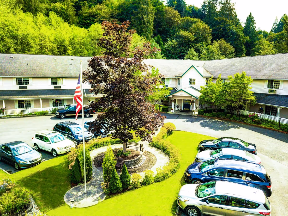 Exterior view of Cascade Valley Senior Living facility showing a two-story building with a gray roof surrounded by lush green trees. In front of the building is a circular driveway with several parked cars and a landscaped roundabout featuring a tree, benches, and an American flag on a flagpole.