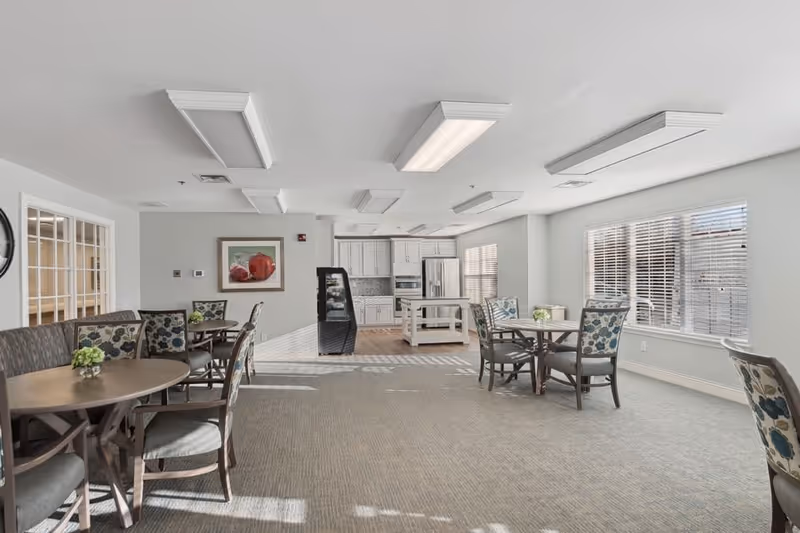 A bright and spacious common area in a senior living facility featuring multiple round tables with floral-patterned chairs, a small kitchen area with white cabinets, a refrigerator, and an island. Large windows with blinds allow natural light to fill the room.