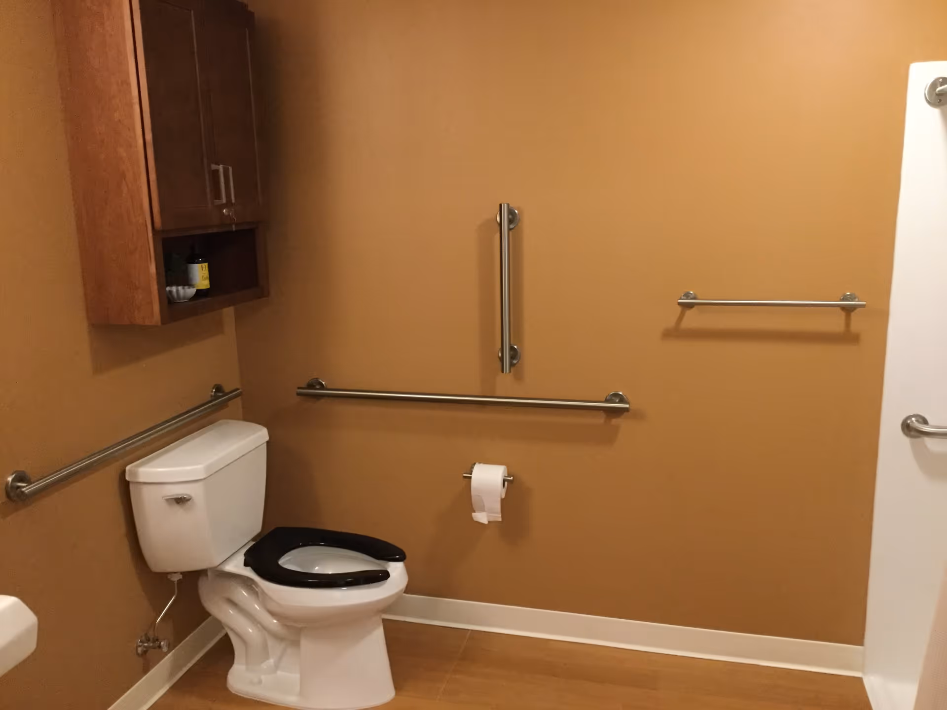 A bathroom with tan walls and a white toilet with a black seat. There are multiple stainless steel grab bars on the walls around the toilet for accessibility. A wooden cabinet is mounted on the wall above the toilet, and a toilet paper holder is mounted on the wall next to the toilet.