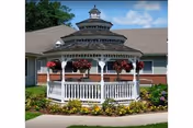 A white wooden gazebo with a multi-tiered roof and hanging flower baskets, situated in a garden area with green grass, colorful flowers, and a paved walkway. The background shows part of a building with a sloped roof under a blue sky with some clouds.