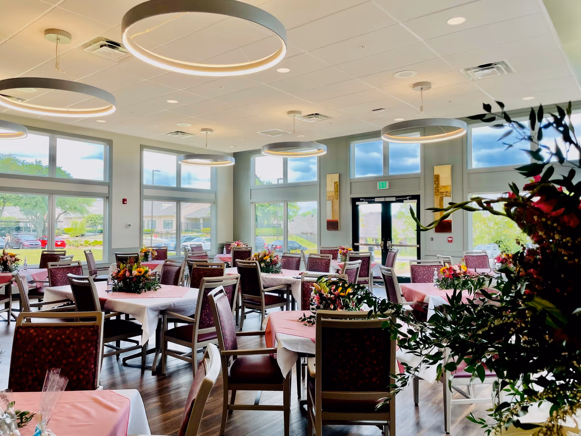 A bright dining room with multiple tables covered in pink tablecloths and white overlays, each adorned with colorful floral centerpieces. The room has large windows letting in natural light, modern circular ceiling lights, and wooden chairs with patterned upholstery. Outside the windows, cars and greenery are visible.