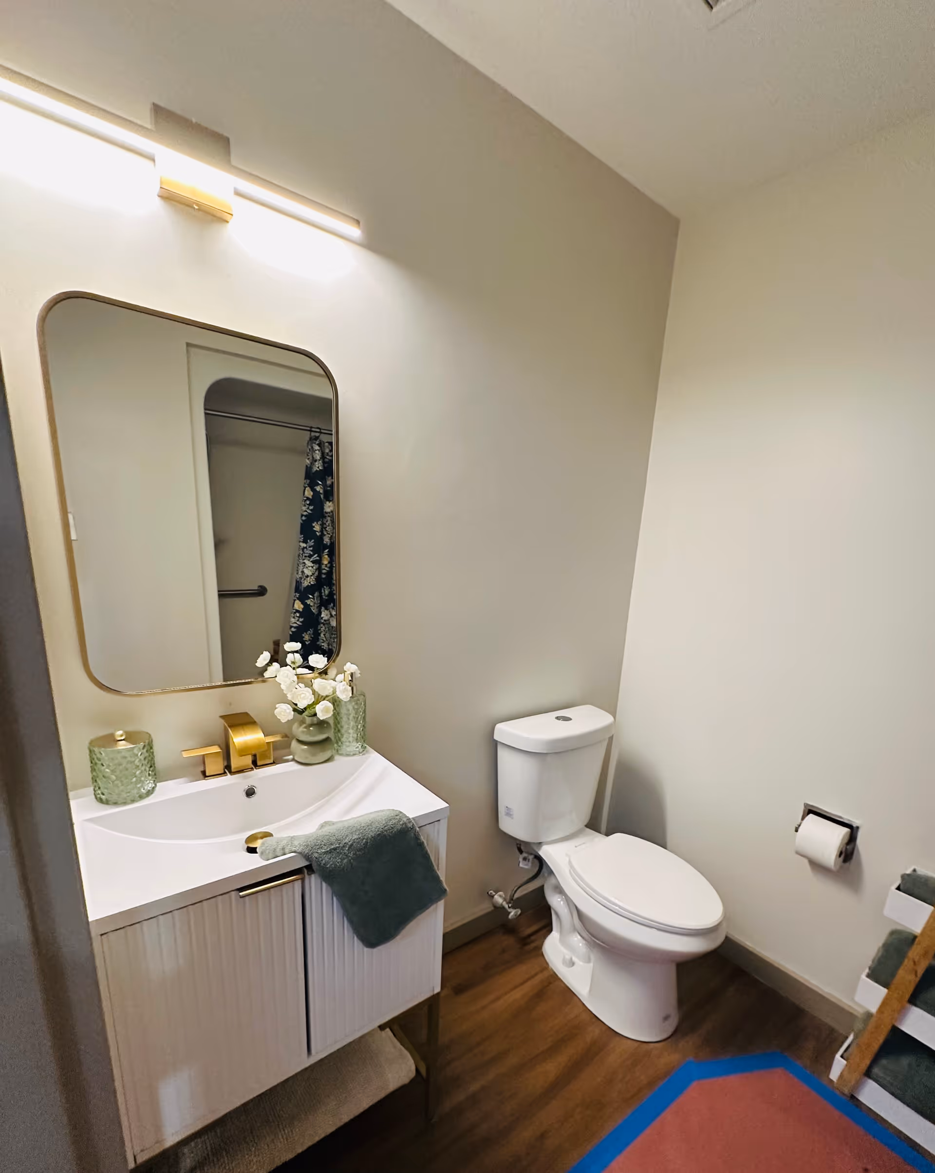 A clean and modern bathroom with a white toilet and a white vanity with a gold faucet. Above the vanity is a rectangular mirror with rounded corners and a light fixture. There are decorative items including a green candle holder, a small vase with white flowers, and a green towel hanging on the vanity. The floor is wood, and a shower curtain with a floral pattern is partially visible in the mirror reflection.
