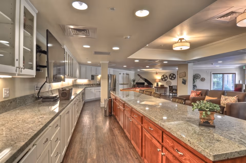 A spacious communal kitchen and lounge with a long granite island, cabinetry, TV, and seating in a senior living facility.