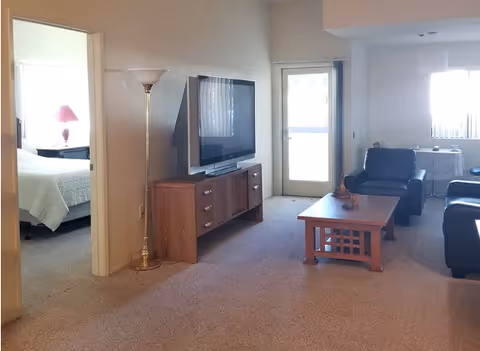 Interior view of a senior living facility showing a living room with a flat-screen TV on a wooden stand, a floor lamp, a wooden coffee table, and black leather chairs. To the left, there is a bedroom with a bed and a bedside table with a lamp. In the background, there is a dining area with a table and chairs near a window and a door leading outside.