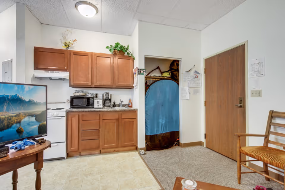 Compact kitchenette and living area with wooden cabinets, a small stove and microwave, a TV on a table, and an entry door with a closet.