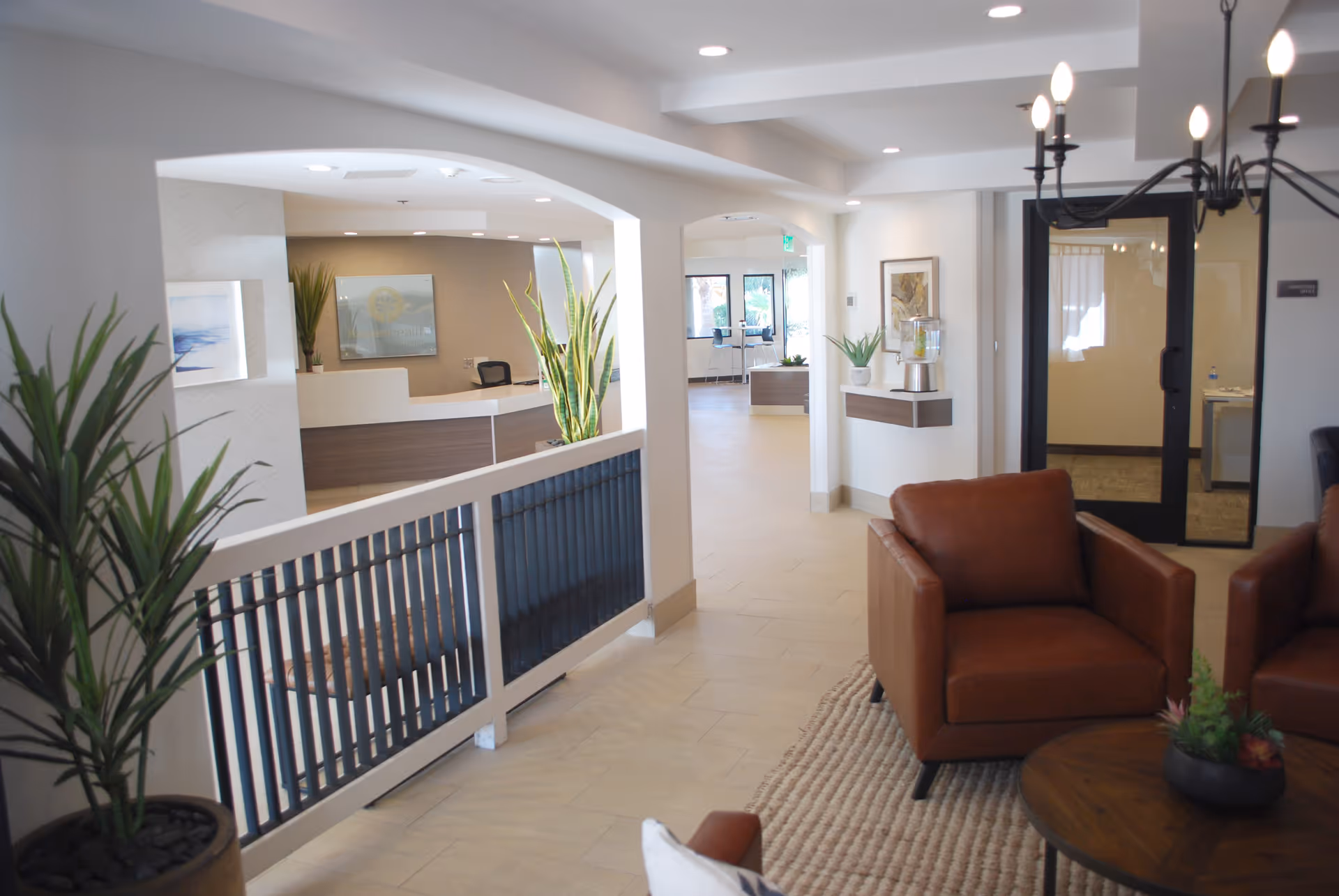 Bright senior living lobby with leather armchairs, a reception desk, potted plants, and a hallway leading to other rooms.