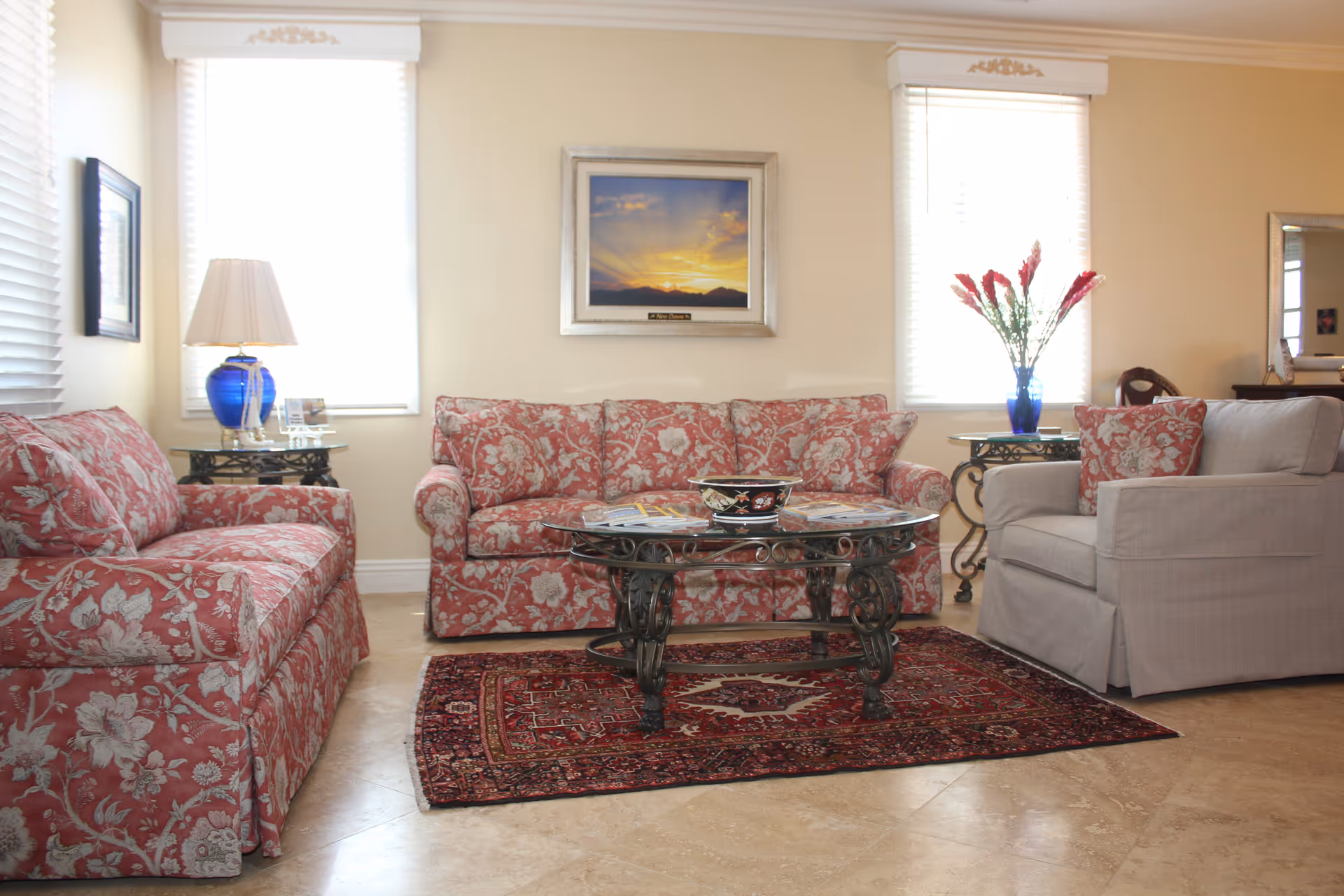 A bright living room with two floral patterned red sofas and one light gray armchair arranged around a glass coffee table with ornate black metal legs. The floor has a red patterned rug, and there are two windows with white blinds letting in natural light. A blue vase with red flowers sits on a side table next to the armchair, and a blue lamp is on a side table next to one of the sofas. A framed picture of a sunset hangs on the wall between the windows.