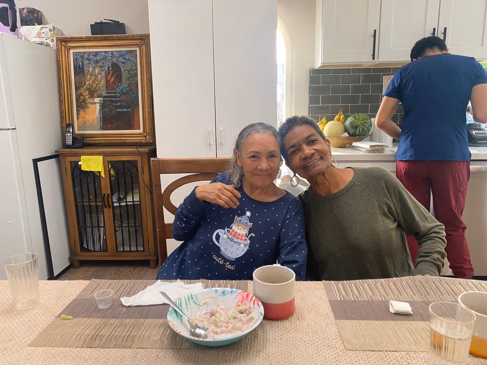 Two women sit together at a dining table with bowls and cups, while a staff member works in the kitchen behind them.