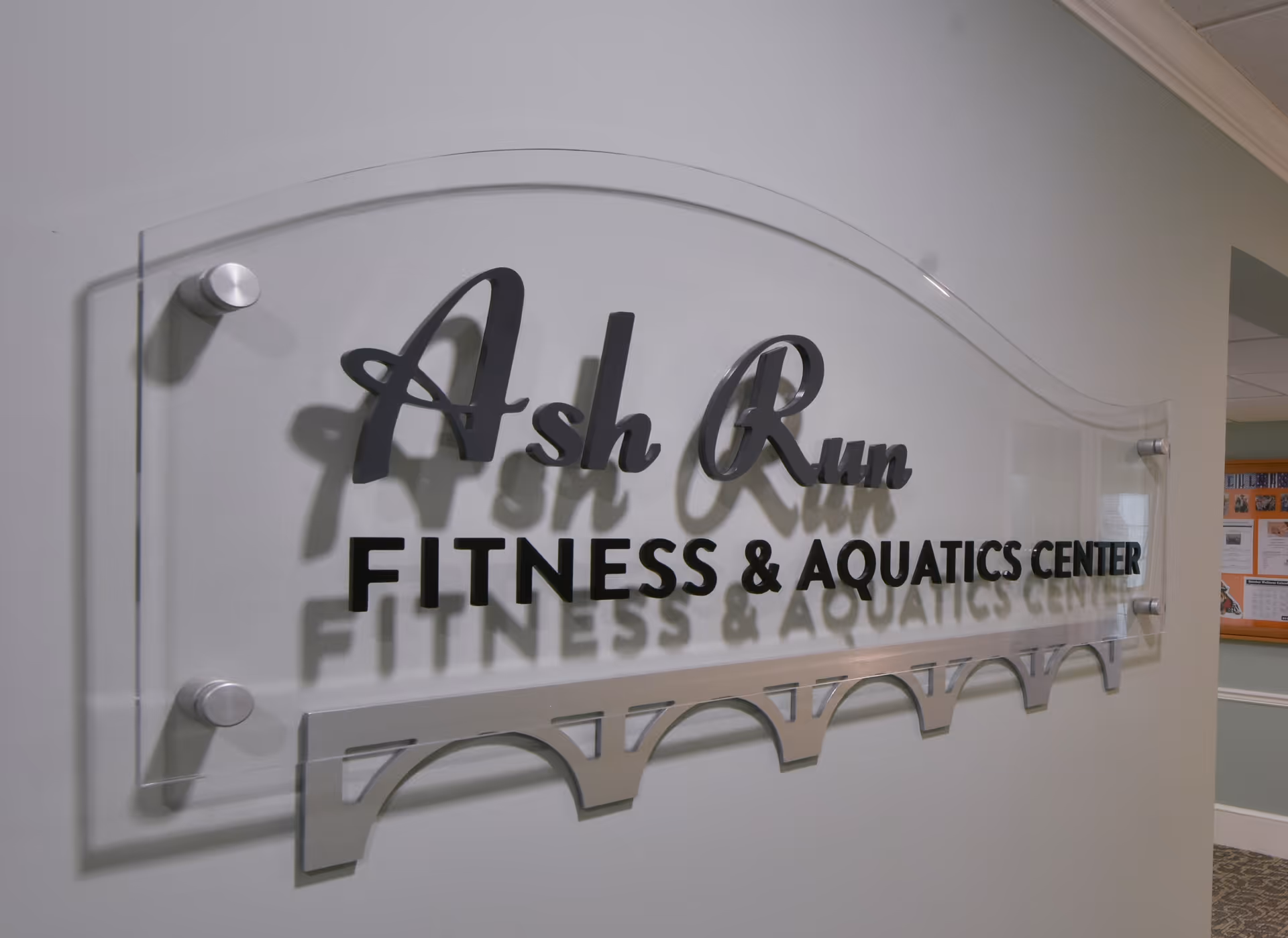 A clear acrylic wall sign with black lettering that reads 'Ash Run FITNESS & AQUATICS CENTER' mounted on a light gray wall in an indoor hallway.