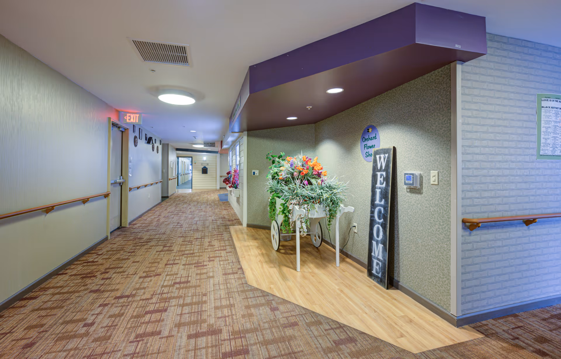 A well-lit hallway in a senior living facility with patterned carpet and handrails on both sides. On the right side, there is a small area with wooden flooring featuring a decorative white cart filled with colorful artificial flowers and a vertical wooden sign that reads 'WELCOME'. Above the cart, a round sign says 'Orchard Flower Shop'. The walls are decorated with light-colored wallpaper and there is an exit door with a red exit sign on the left side of the hallway.