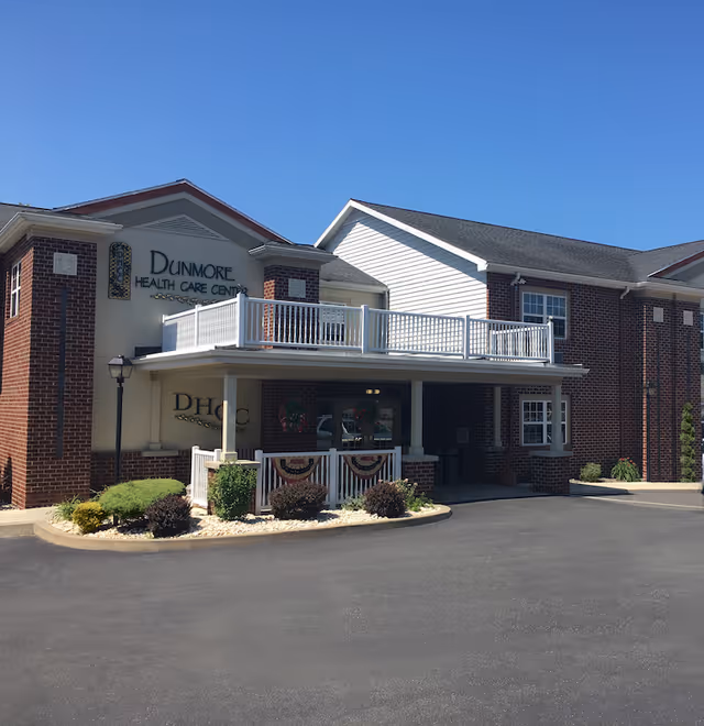 Exterior front view of Dunmore Health Care Center, a two-story brick building with white trim and a covered entrance. The building has a balcony with white railings above the entrance and landscaping with bushes and flowers in front. The sky is clear and blue.