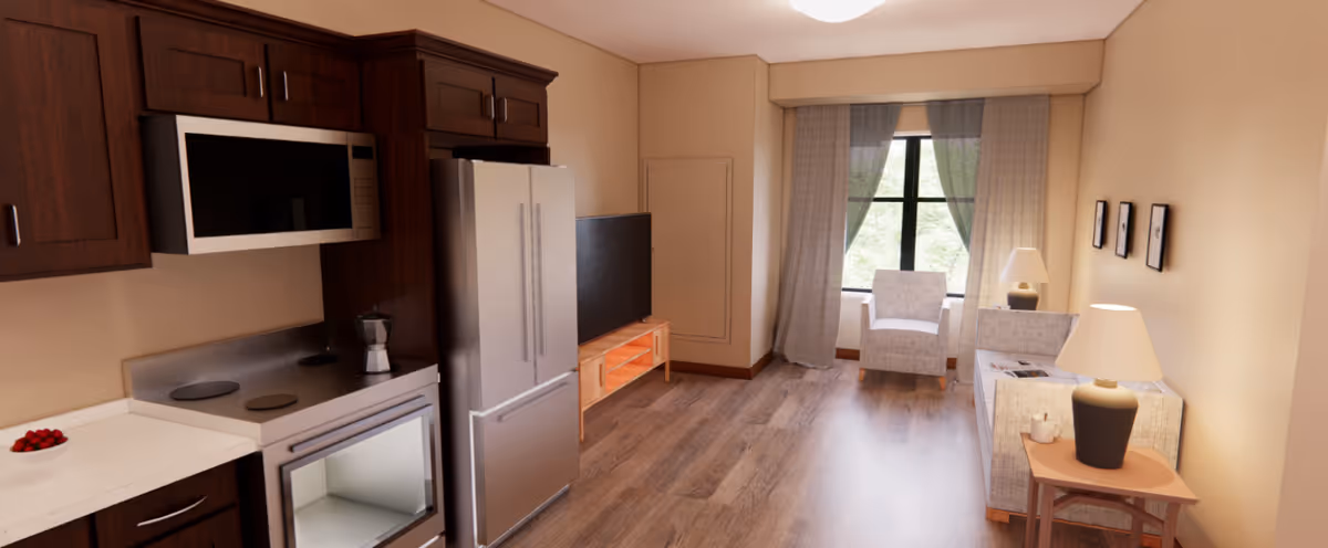 Open studio interior showing a kitchenette with stainless appliances on the left and a living area with chairs, sofa, TV, and window ahead.