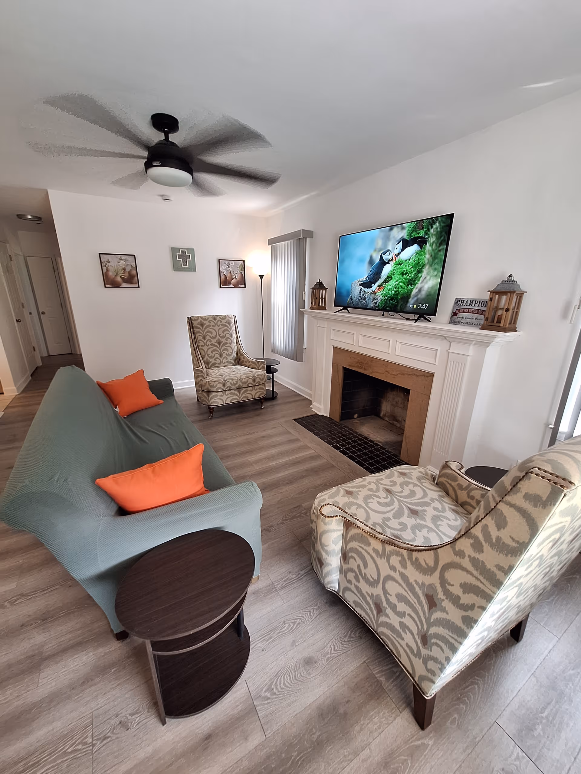 A cozy living room with a green sofa covered with a slipcover and two patterned armchairs. There are two orange pillows on the sofa. A round dark wooden side table is next to the sofa. A white fireplace with a mounted flat-screen TV above it is on the right wall. The TV displays an image of puffins on a mossy rock. Two lantern-style decorations and a small sign are on the fireplace mantel. The room has light wood flooring, white walls, a ceiling fan, and a tall floor lamp near the window with vertical blinds. Two framed pictures and a decorative cross hang on the far wall.