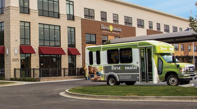 Exterior view of a senior living facility named First & Main Assisted Living and Memory Care, showing a multi-story building with large windows and red awnings. A green and white shuttle bus with the First & Main logo is parked in front of the building on a curved driveway.
