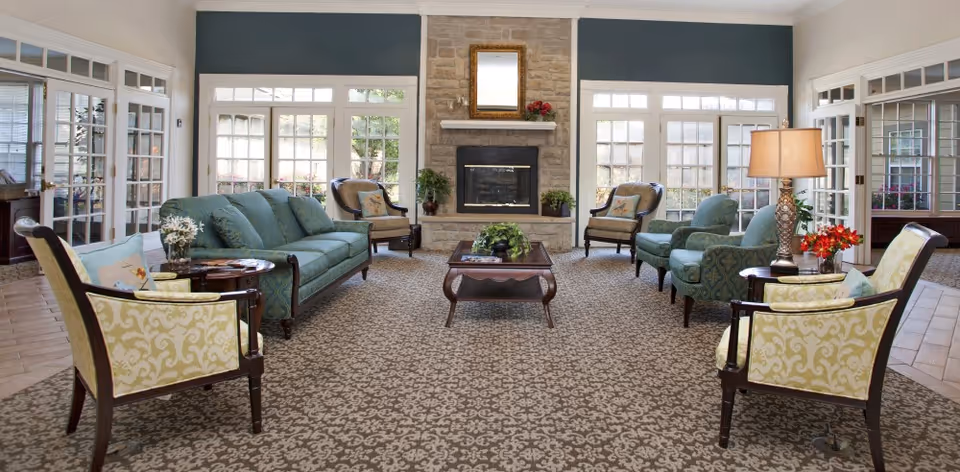 A spacious and well-lit living room with a patterned carpet, a stone fireplace with a mirror above it, and multiple seating options including a green sofa, two green armchairs, and two yellow patterned chairs. There are side tables with lamps and flowers, and large windows letting in natural light.