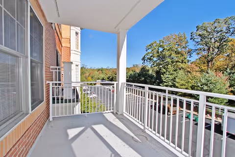 View from a covered balcony with white metal railings overlooking a parking lot and trees with autumn foliage under a clear blue sky.