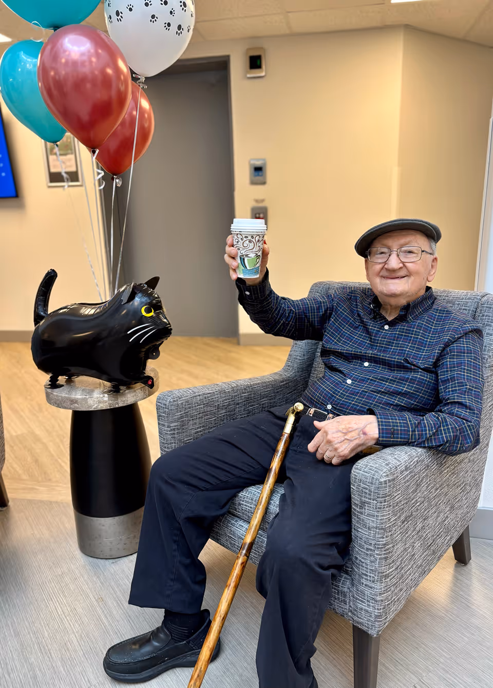 An elderly man wearing a flat cap and glasses sits in a gray armchair, smiling and holding up a decorated paper coffee cup. Next to him is a small round table with a black cat-shaped balloon and several colorful balloons tied to it. The setting appears to be a bright indoor common area with light-colored walls and flooring.