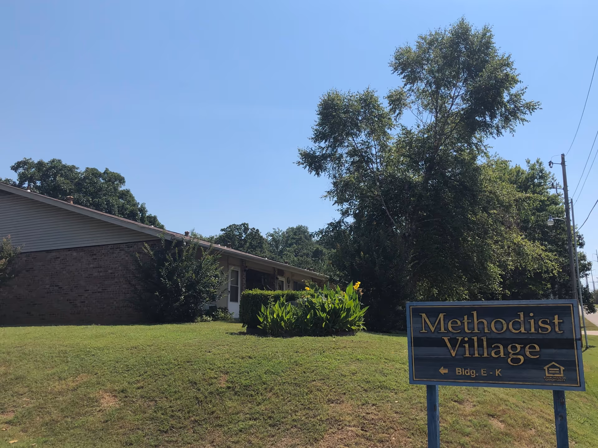 Single-story brick building and lawn with a 'Methodist Village' sign in the foreground under a clear blue sky.