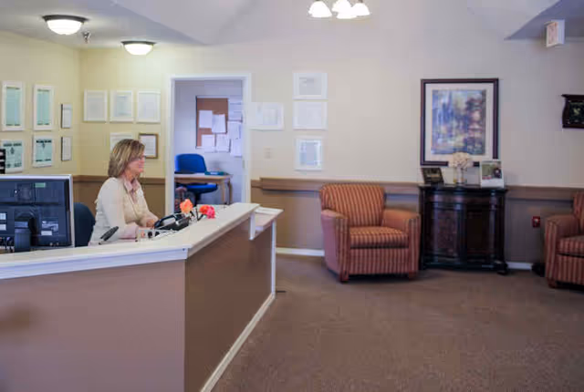 Reception area in a senior living facility with a woman sitting behind a front desk with computer monitors. The room has beige walls, framed pictures, two striped armchairs, a dark wooden cabinet with decorative items, and a doorway leading to another room with a blue chair.