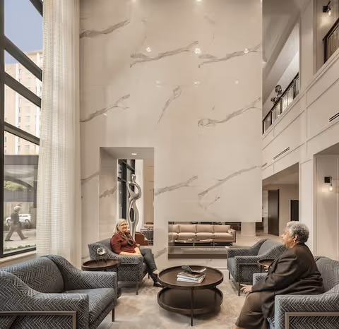 A spacious and elegant living room area in a senior living facility with high ceilings and large windows. Two elderly women are seated on patterned armchairs facing each other, with a round coffee table between them. The room features a tall marble accent wall, modern sculpture, and neutral-toned furniture, creating a warm and inviting atmosphere.