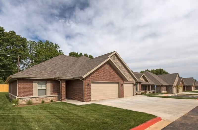 Exterior view of a single-story brick residential building with a two-car garage, a well-maintained lawn, and a driveway under a partly cloudy sky.