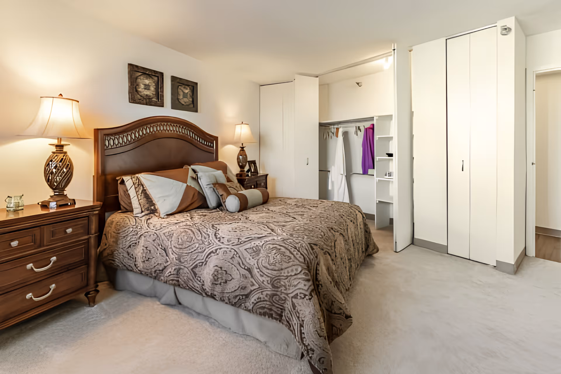 A cozy bedroom featuring a large bed with a patterned brown and beige bedspread and multiple pillows. There are two wooden nightstands on either side of the bed, each with a decorative lamp. The room has beige carpeting and white walls with two framed artworks above the bed. An open closet with white doors reveals hanging robes and shelves. A doorway leads to another room.