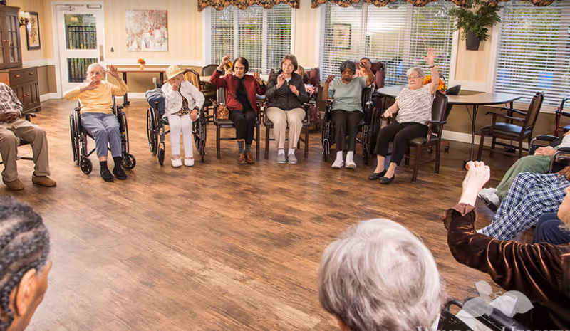 A group of elderly individuals sitting in a circle in a well-lit room with wooden flooring, some in wheelchairs and others in chairs, raising their hands as if participating in an activity or exercise. The room has large windows with blinds, plants, and framed artwork on the walls.