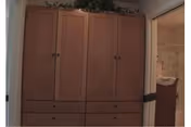 A tall wooden armoire with multiple drawers and cabinets topped with greenery, positioned next to a bathroom doorway.