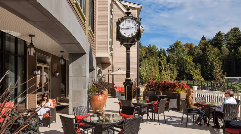 Outdoor patio area at Aegis Living Queen Anne Rodgers Park with several round glass tables and wicker chairs with red cushions. A large decorative clock on a black post stands near the building. Three people are seated at two of the tables enjoying the sunny day. The background shows green trees and a clear blue sky.