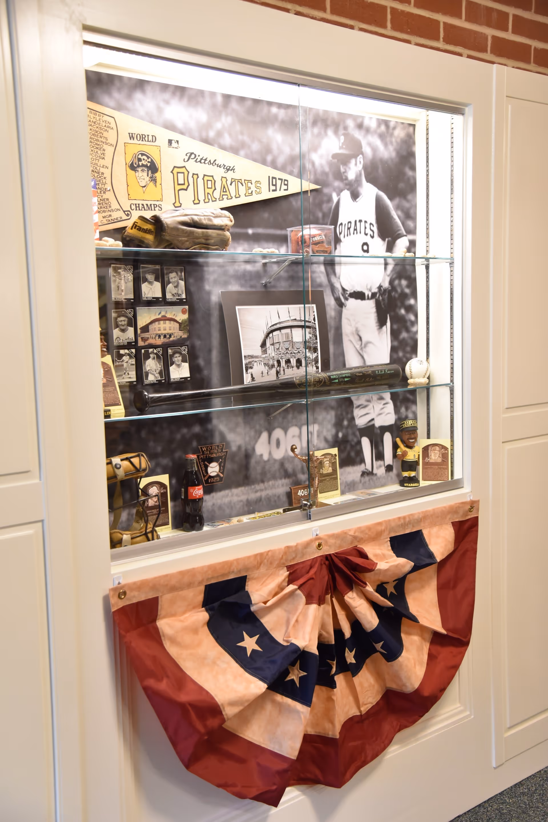 A glass display case featuring Pittsburgh Pirates memorabilia including a 1979 World Champs pennant, a baseball glove, vintage photos, a baseball bat, a baseball, a Coca-Cola bottle, and a bobblehead figure. The background has a large black and white photo of a Pirates player. Below the display case is a red, white, and blue patriotic bunting decoration.