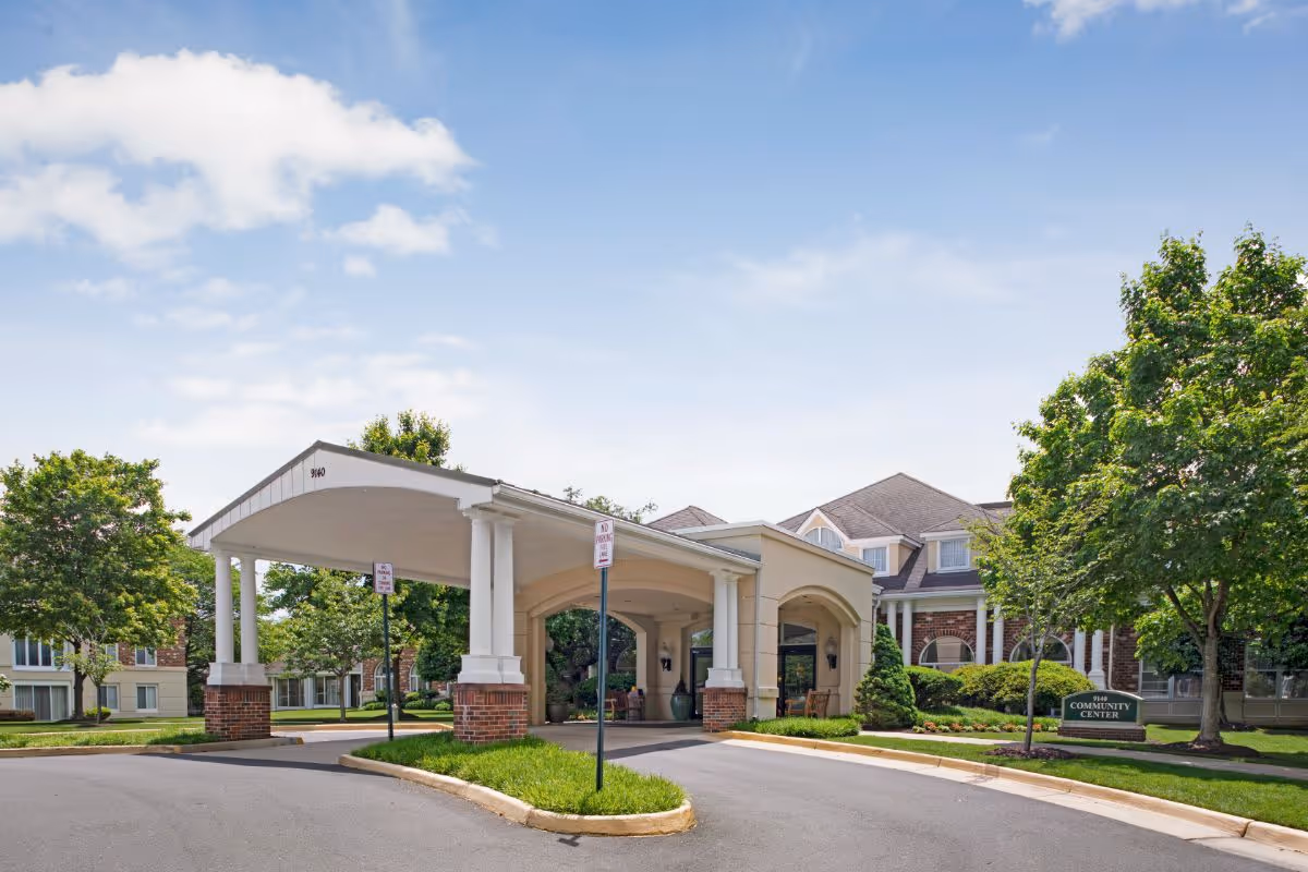 Exterior view of The Fairfax at Belvoir Woods community center entrance with a covered driveway, surrounded by green trees and well-maintained landscaping under a partly cloudy sky.