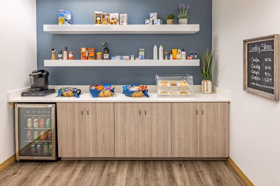 A small snack and beverage area with a mini fridge stocked with canned drinks, a coffee maker, three baskets filled with assorted snacks, and a clear display case containing baked goods. Two white floating shelves on a blue-gray wall hold various packaged food items and personal care products. A chalkboard on the right wall lists prices for soda, candy, chips, and cookies under the heading 'Country Store'.