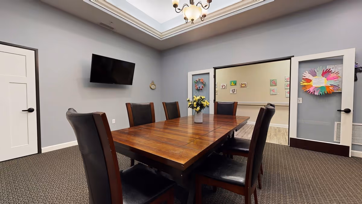 A meeting or dining room with a large wooden table surrounded by six dark leather chairs. The walls are painted light gray with white trim, and a flat-screen TV is mounted on one wall. There are two glass doors leading to another room decorated with colorful artwork. A vase with yellow flowers is placed in the center of the table.