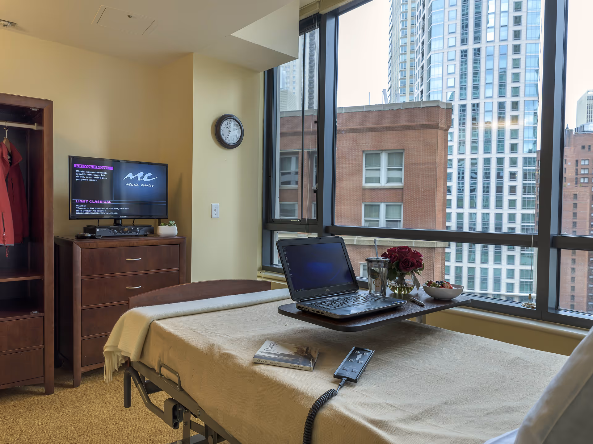 A senior living bedroom with an adjustable bed, overbed laptop tray, dresser and large windows overlooking city buildings.