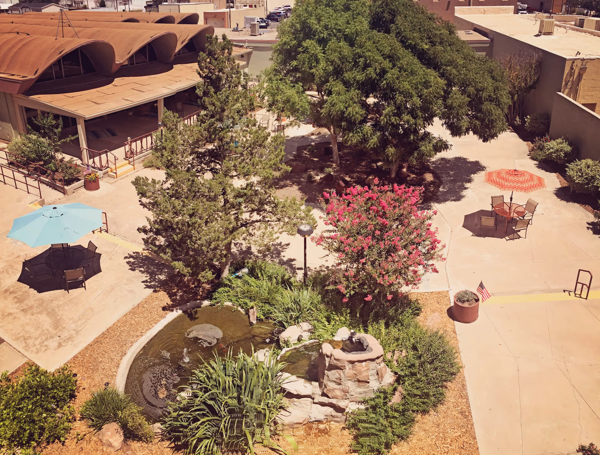 Outdoor courtyard area with trees, flowering bushes, a small pond with rocks, and two seating areas with tables and umbrellas, one blue and one red. Surrounding buildings and a parking lot are visible in the background.