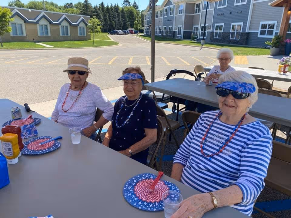 Four elderly women sitting at outdoor tables under a canopy, enjoying a sunny day. They are wearing casual summer clothes, sunglasses, and patriotic-themed accessories. Behind them are senior living apartment buildings and a parking lot.
