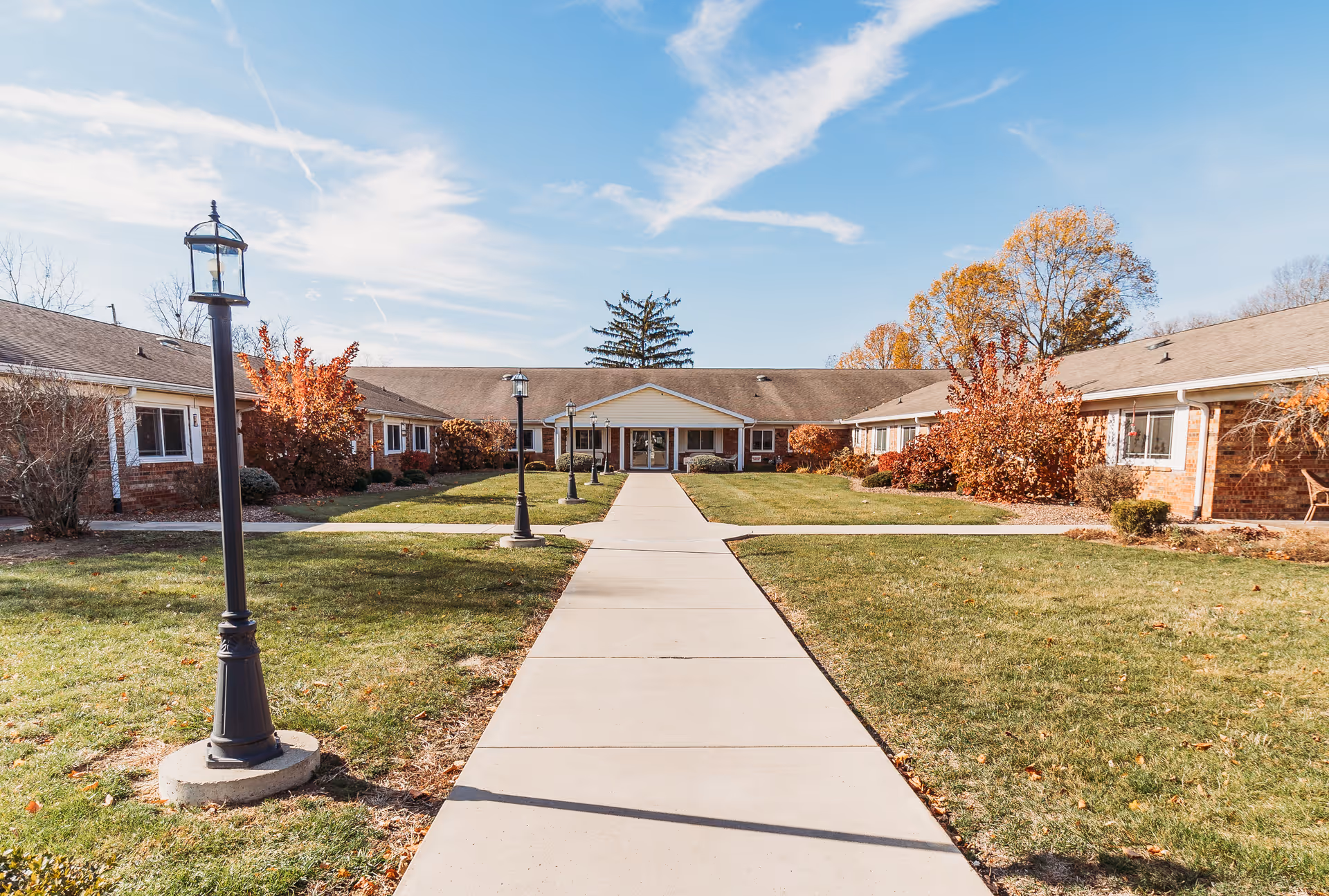 A wide concrete walkway leads through a grassy courtyard with lamp posts on either side, surrounded by a single-story brick building with white trim and a peaked entrance. Trees with autumn foliage and clear blue sky are visible in the background.