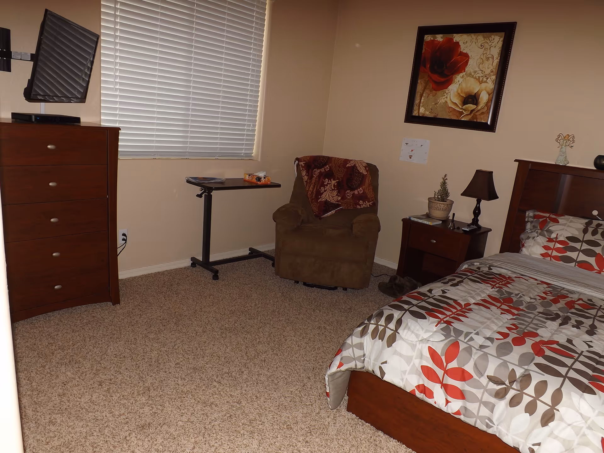 A bedroom with a bed covered in a patterned comforter with red, gray, and beige leaves. There is a wooden nightstand with a lamp and a small plant, a brown armchair with a blanket draped over it, a small rolling table, a wooden dresser, and a window with closed blinds. A framed floral painting hangs on the wall above the armchair.