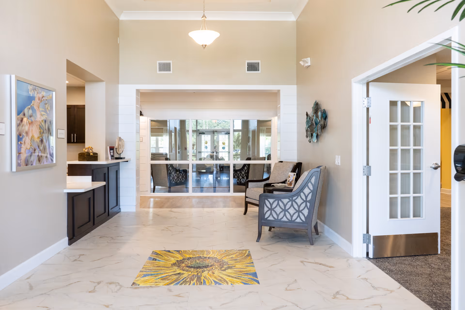 Bright and spacious interior hallway with beige walls and marble-patterned floor tiles featuring a large sunflower design in the center. Two cushioned chairs with patterned backs are placed against the right wall, next to a decorative wall hanging. A white door with glass panels is open on the right side, and a counter with dark wood cabinetry is on the left. Large windows and glass doors are visible at the far end, allowing natural light to fill the space.