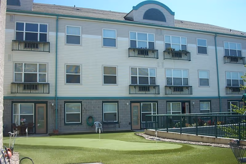 Exterior view of a three-story residential building with multiple windows and small balconies. The ground level has doors leading to a green artificial turf area with a putting green and some outdoor chairs and tables. The building has light-colored siding with a stone base and green trim.