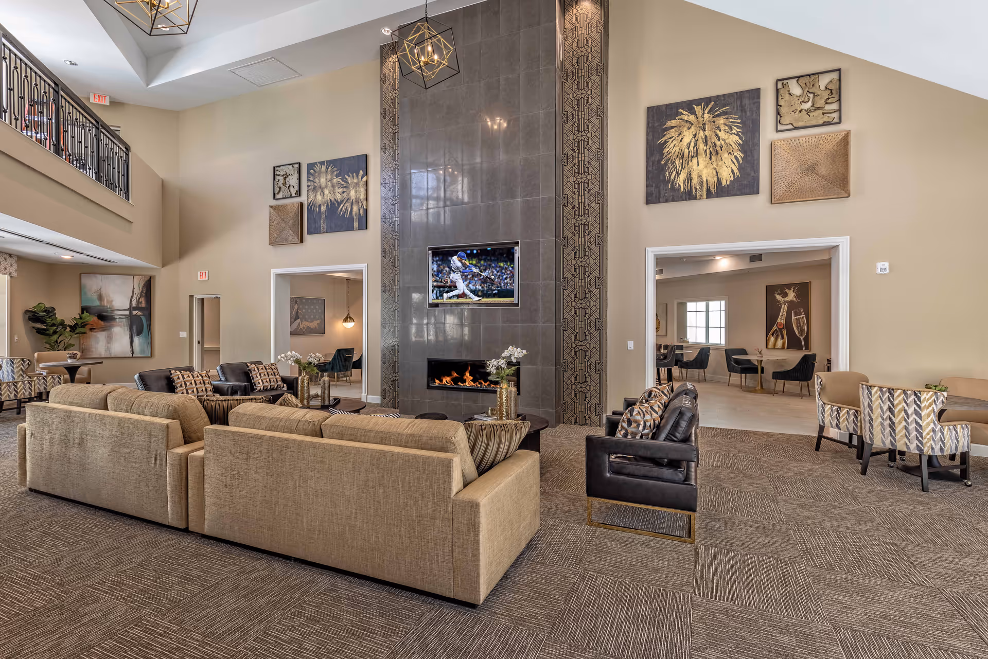 Spacious senior living facility common area with beige and black sofas arranged around a modern fireplace with a TV mounted above it. The room features high ceilings, decorative wall art, and pendant lighting. Adjacent rooms with additional seating and artwork are visible through large doorways.