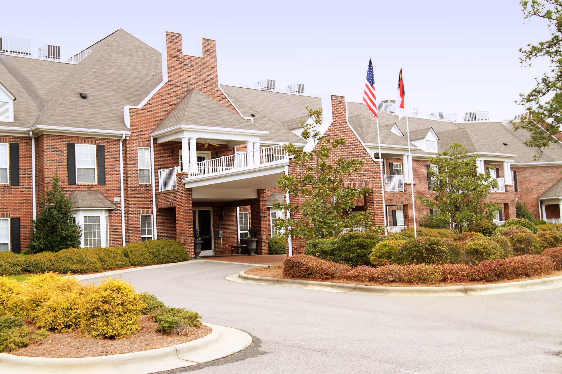 Exterior view of a large brick senior living facility building with multiple windows, a covered entrance with white columns, two flagpoles displaying the American and North Carolina flags, and landscaped bushes and trees around the driveway.